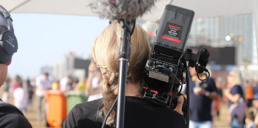 Auf dem Bild ist eine Frau mit einer Filmkamera auf der Schulter von hinten zu sehen