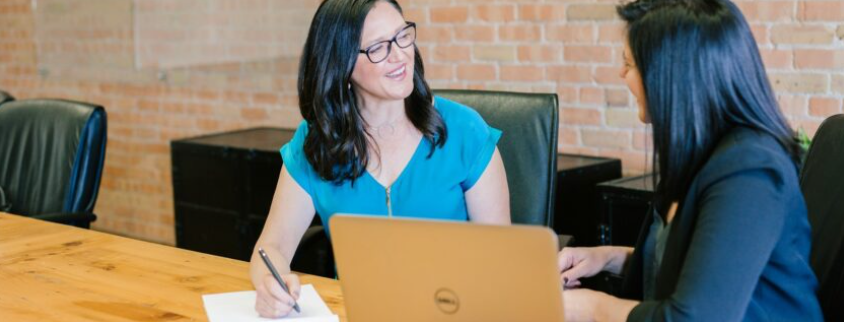 Auf dem Bild sind zwei Frauen zu sehen, die sich an einem Tisch unterhalten