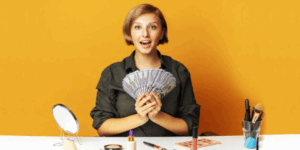 Junge Frau mit vielen Geldscheinen in der Hand sitzt am Tisch. Darauf viel Kosmetik-Artikel.
